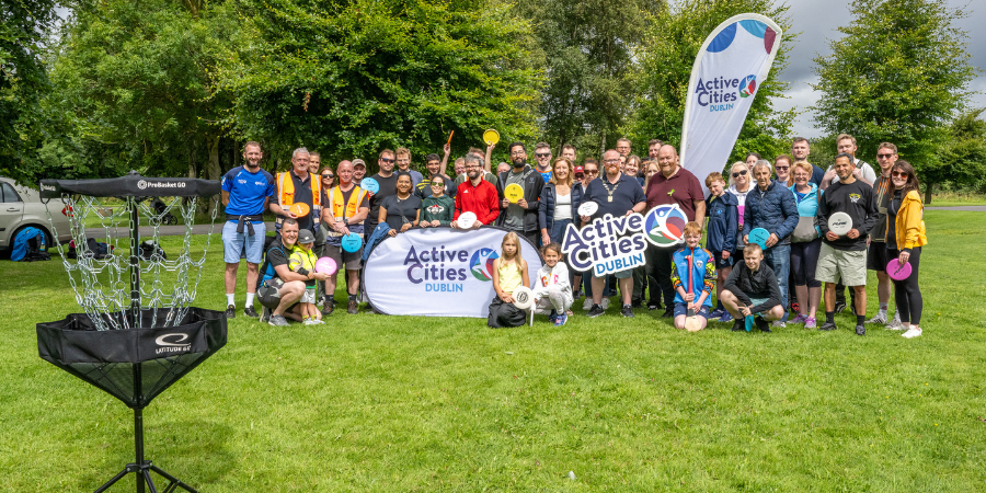 Grúpa mór daoine de gach aois agus cúlra ag seasamh le chéile i bpáirc do lá imeartha disc golf eagraithe ag Active Cities Dublin. Tá na rannpháirtithe ag coinneáil dioscaí ildaite agus ag miongháire os comhair meirgí agus bratacha branda. Tá ciseán sprice disc golf le feiceáil sa tulra ar an bhféar glas. Tá atmaisféar cairdiúil agus fuinniúil ann, ag ceiliúradh spórt pobail agus gníomhaíocht fhisiciúil chuimsitheach faoin aer.